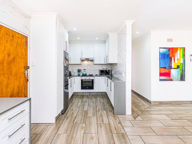 Modern kitchen featuring white cabinetry, stone countertops, integrated oven and hob, and wood-look tiled flooring, seamlessly connected to the adjacent living area.
