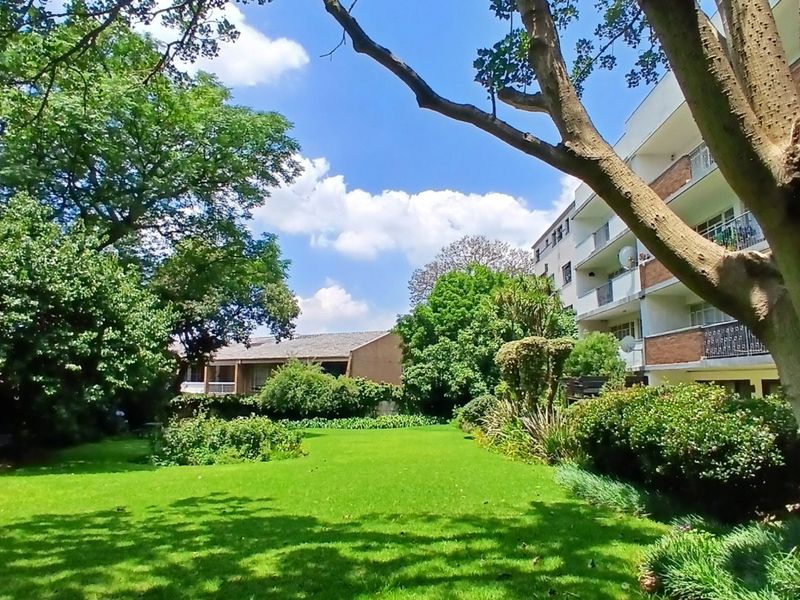 A well-maintained communal garden area featuring a lush green lawn, mature trees, and views of the apartment building's exterior facade and balconies.