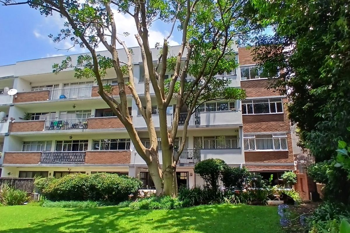 Rear view of a multi-story apartment building featuring brick facade, private balconies, and a well-maintained communal garden with mature trees.