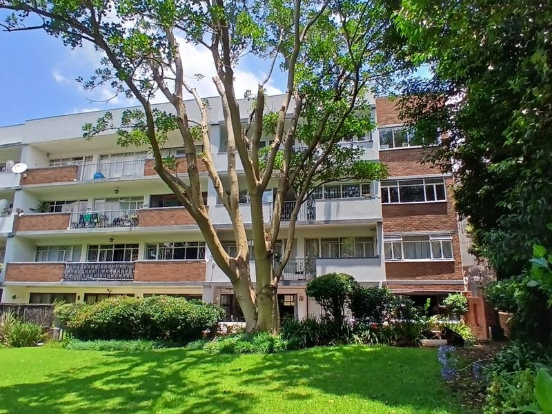 Rear view of a multi-story apartment building featuring brick facade, private balconies, and a well-maintained communal garden with mature trees.