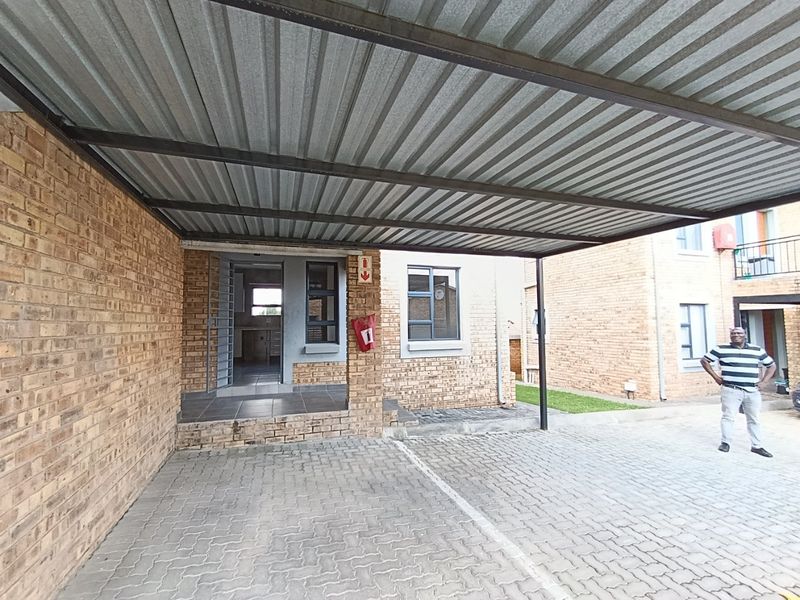 A covered carport area with a corrugated metal roof, brick exterior walls, and paved driveway, providing sheltered parking for the apartment unit.