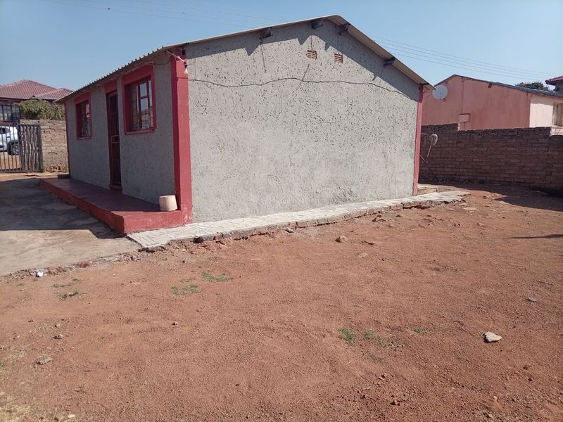 Rear view of a detached house featuring a textured plaster finish, metal roofing, and a surrounding dirt yard with a brick boundary wall.