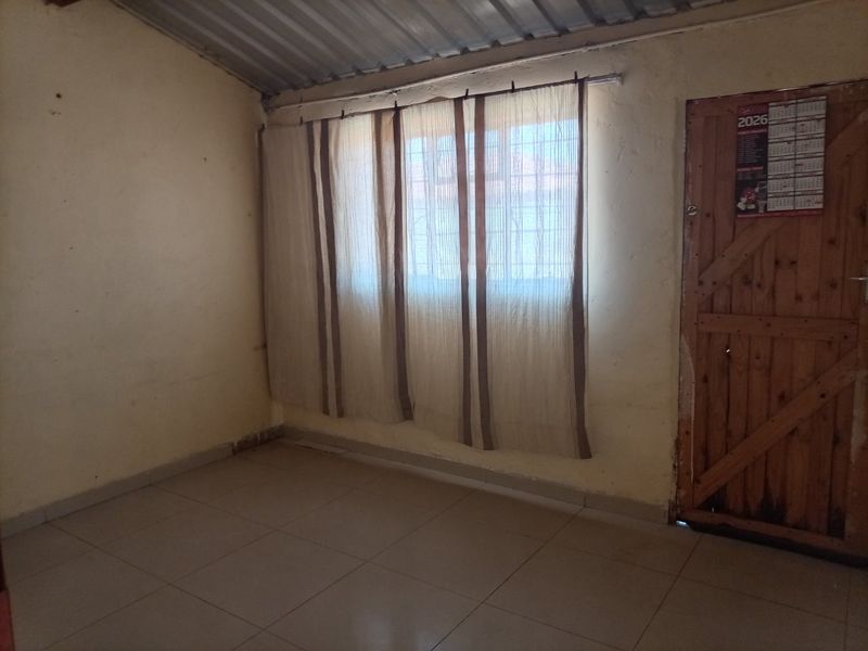 An empty bedroom featuring tiled flooring, exposed metal roof sheeting, and a timber door with security bars on the window.