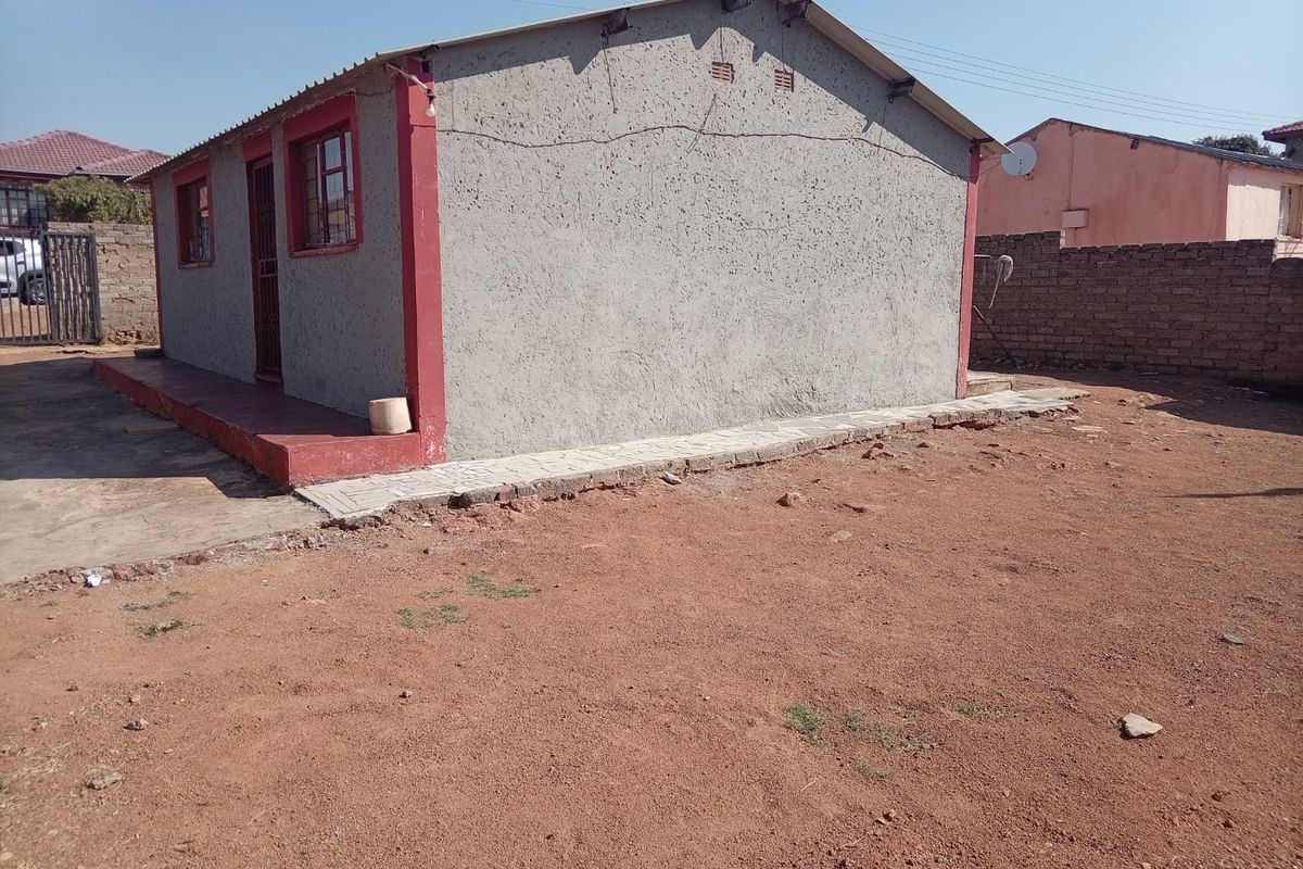 Rear view of a detached house featuring a textured plaster finish, metal roofing, and a surrounding dirt yard with a brick boundary wall.