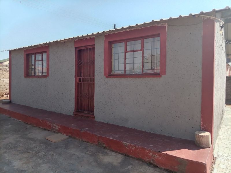 Front elevation of a single-story dwelling featuring a concrete plinth, security gate, and steel-framed windows with a corrugated metal roof.
