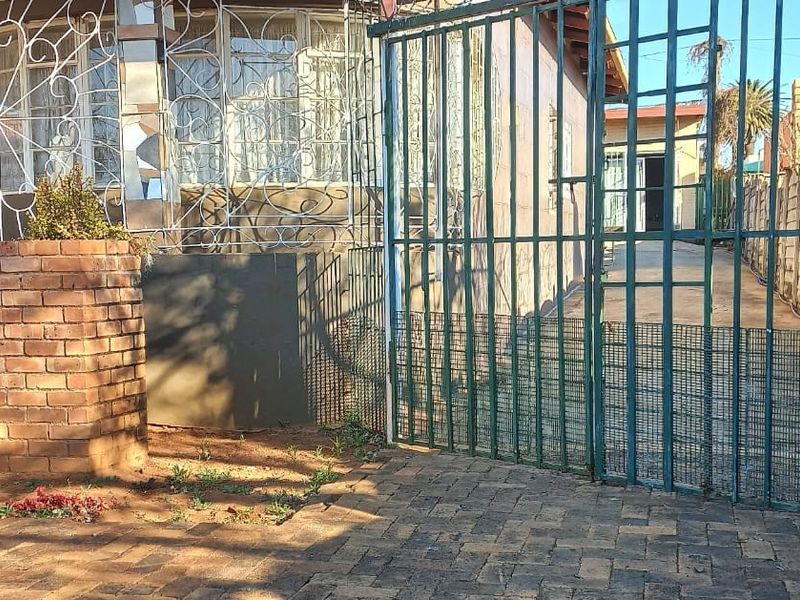 Front exterior view showing a brick driveway, metal security gate, and perimeter walling with security bars on the house windows.