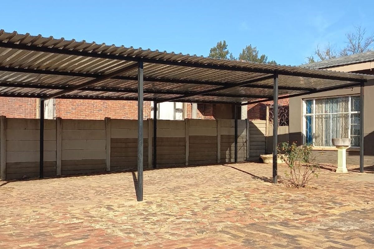 Rear exterior area featuring a large corrugated metal carport, brick paved ground, and precast concrete boundary walling adjacent to the main house structure.