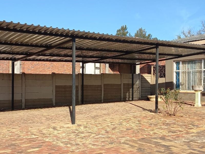 Rear exterior area featuring a large corrugated metal carport, brick paved ground, and precast concrete boundary walling adjacent to the main house structure.