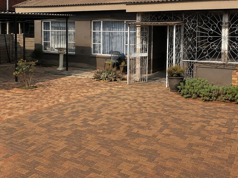 A well-maintained front exterior featuring a spacious paved driveway, a covered carport, and secure metal security gates and window bars.