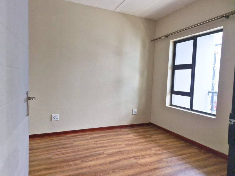 A clean, empty bedroom featuring modern laminate flooring, neutral wall finishes, and an aluminium-framed window providing natural light.