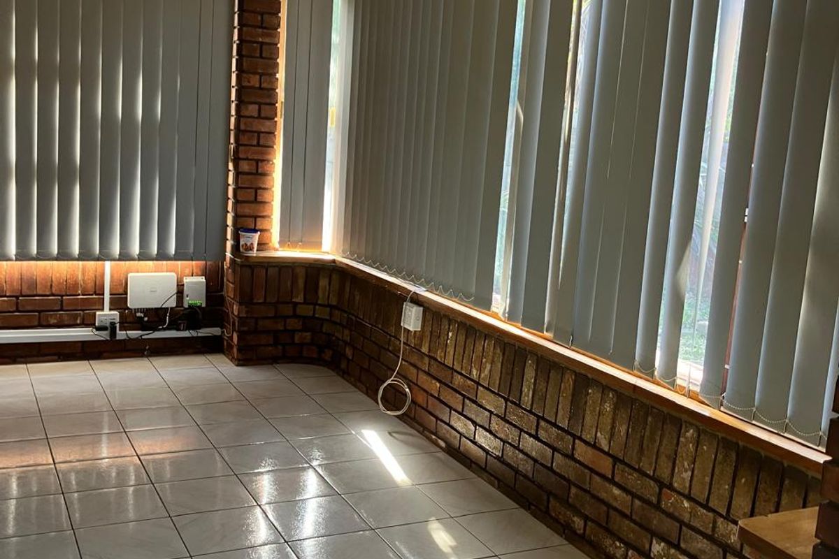 A bright, empty living area featuring exposed brick walls, ceramic floor tiling, and large windows fitted with vertical blinds.