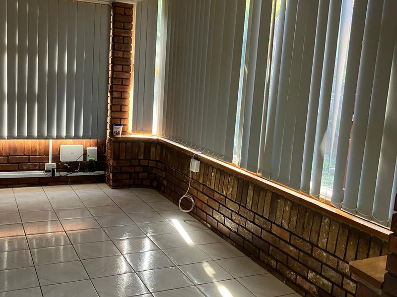 A bright, empty living area featuring exposed brick walls, ceramic floor tiling, and large windows fitted with vertical blinds.