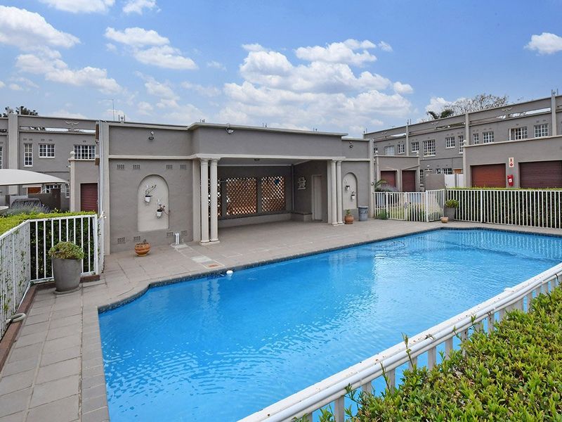 A well-maintained swimming pool area featuring a covered patio with decorative columns, tiled decking, and secure white metal perimeter fencing.