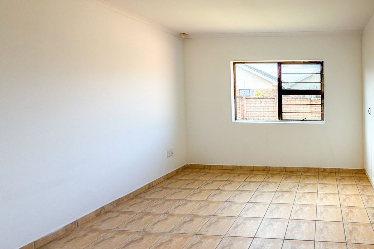 A bright, empty bedroom featuring neutral tiled flooring, white walls, and built-in cupboards with secure window burglar bars.