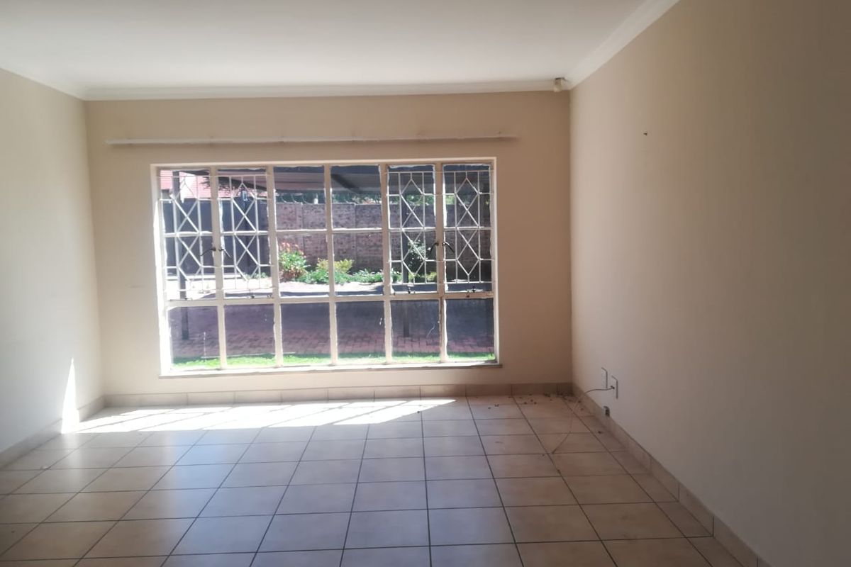 A spacious, empty living room featuring neutral tiled flooring, coving, and large windows fitted with security bars, offering a blank canvas for interior design.