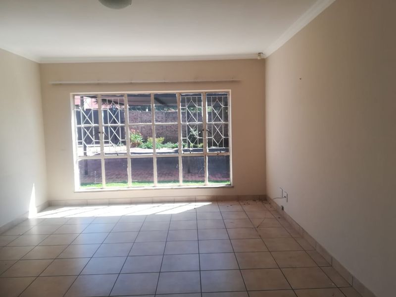 A spacious, empty living room featuring neutral tiled flooring, coving, and large windows fitted with security bars, offering a blank canvas for interior design.