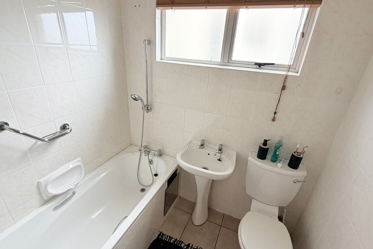 A functional bathroom featuring a bathtub with shower mixer, pedestal basin, toilet, and neutral wall tiling with a frosted window for privacy.
