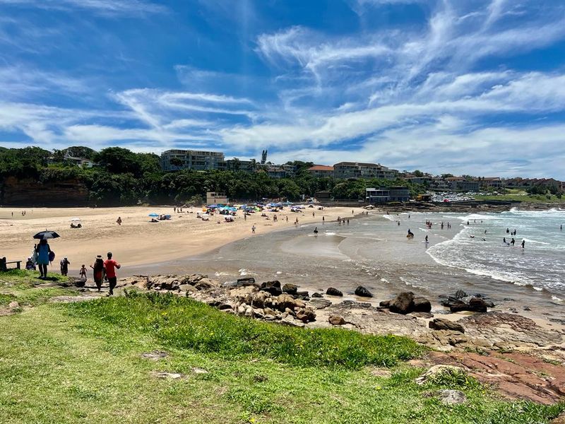 A scenic coastal view overlooking a sandy beach, rocky shoreline, and the surrounding residential neighborhood under a clear blue sky.