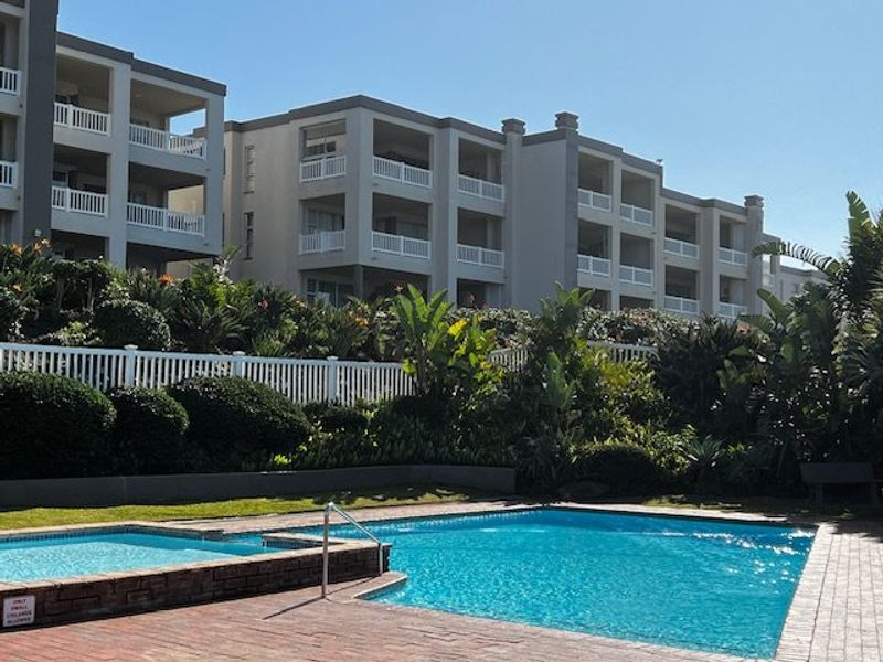 A well-maintained communal swimming pool area featuring paved surrounds, landscaped gardens, and views of the apartment complex balconies.