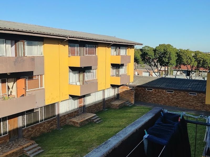 View from a balcony overlooking a communal garden and the rear facade of a multi-story apartment complex with brick and painted finishes.