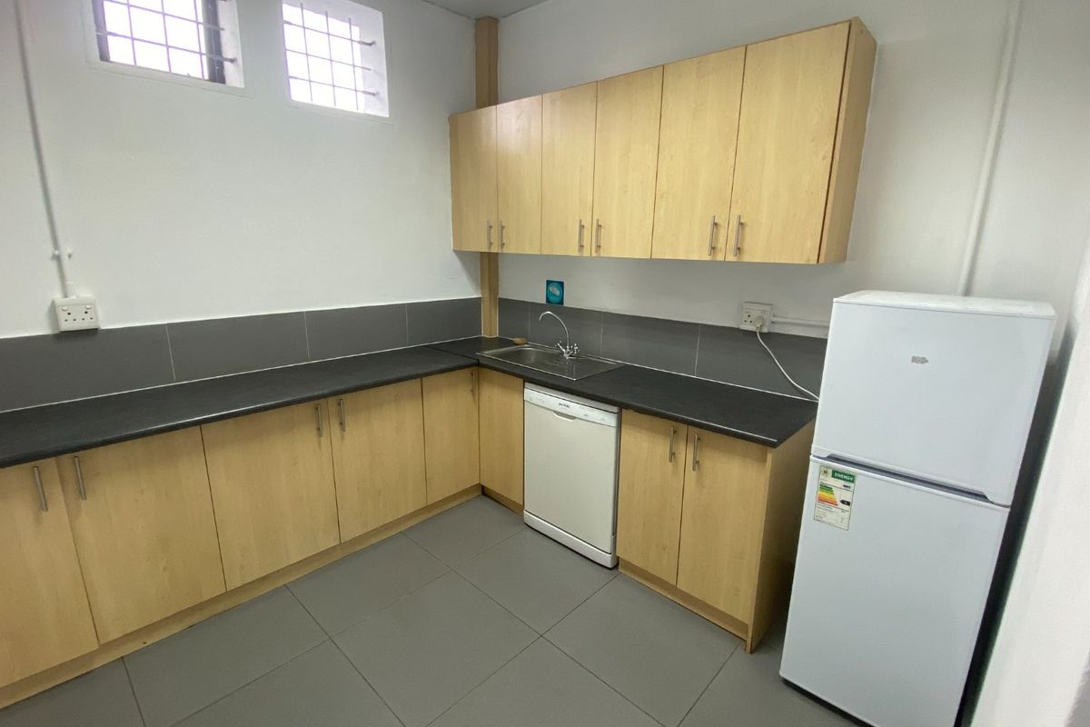A clean, functional kitchenette featuring light wood-finish cabinetry, dark countertops, tiled flooring, and a stainless steel sink with security-barred windows.