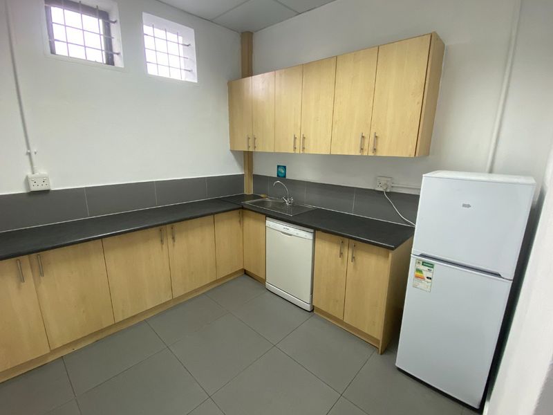 A clean, functional kitchenette featuring light wood-finish cabinetry, dark countertops, tiled flooring, and a stainless steel sink with security-barred windows.