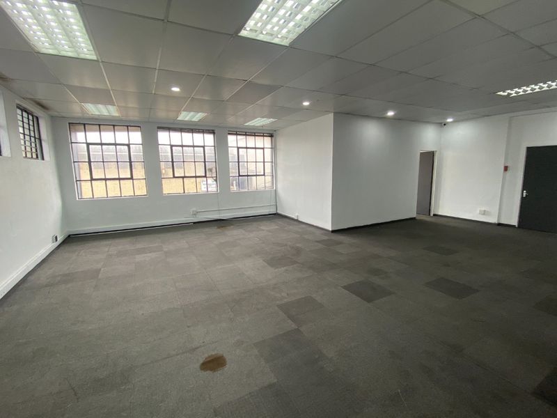 Spacious open-plan office area featuring industrial-style windows, suspended ceilings with recessed lighting, and carpet tile flooring.
