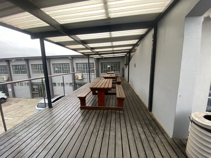 Covered outdoor balcony area featuring timber decking, a corrugated roof structure, and steel cable railings, overlooking the commercial parking area.