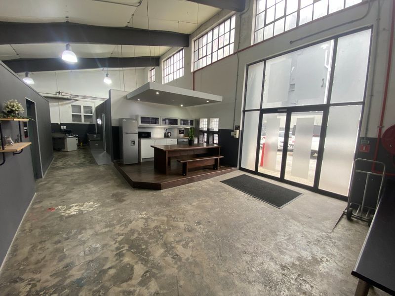 Spacious open-plan commercial area featuring industrial concrete floors, exposed steel beams, large windows, and a raised kitchenette platform with modern cabinetry.
