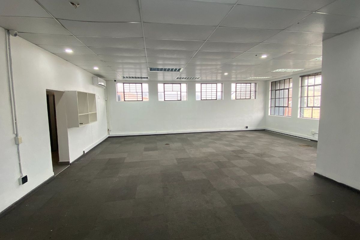 A spacious, empty commercial office area featuring carpet tiles, a suspended ceiling with recessed lighting, and security-barred windows.