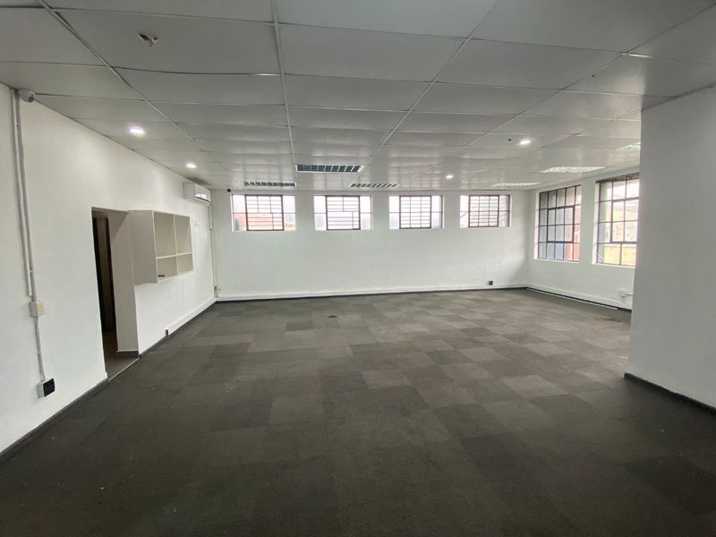 A spacious, empty commercial office area featuring carpet tiles, a suspended ceiling with recessed lighting, and security-barred windows.