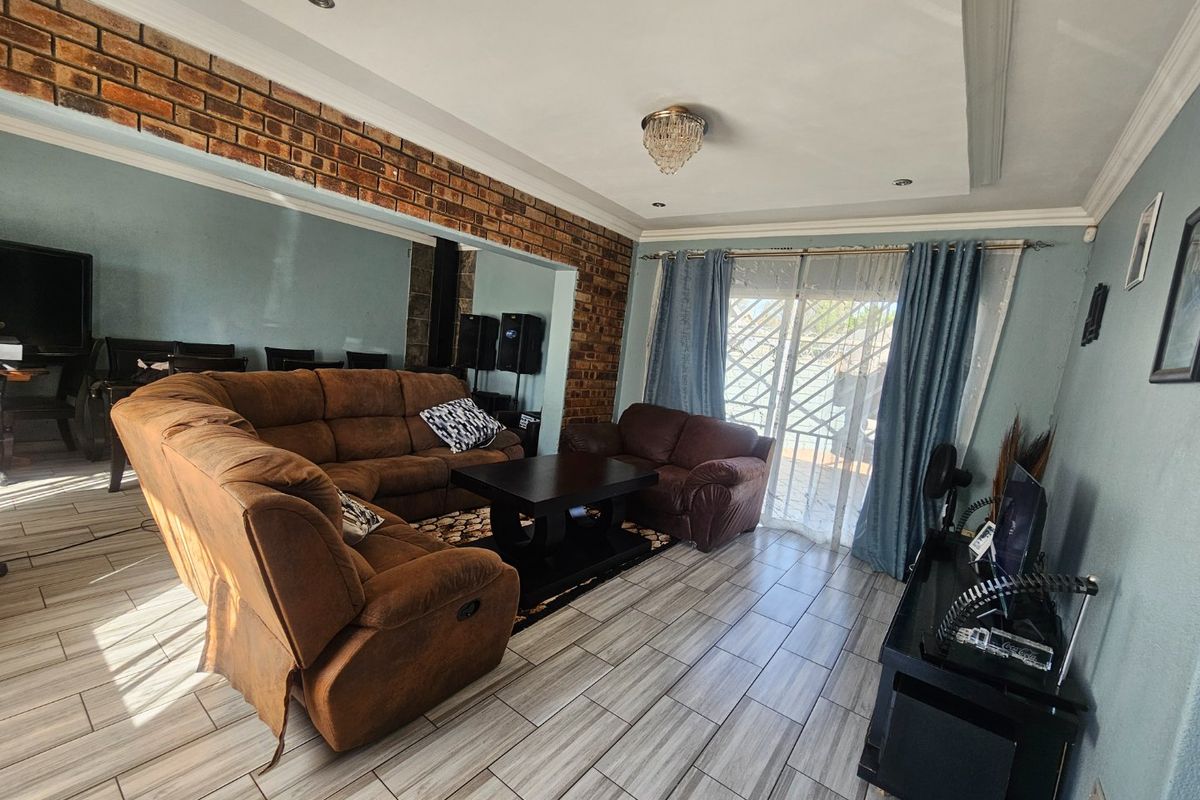 A spacious living room featuring tiled flooring, an exposed brick accent wall, and a sliding glass door with security gate providing access to the exterior.
