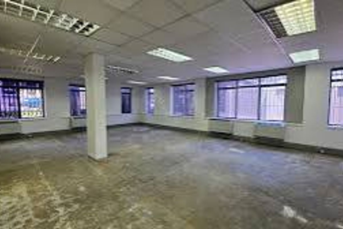 An open-plan commercial office space featuring suspended ceilings, fluorescent lighting, security bars on windows, and exposed concrete flooring.