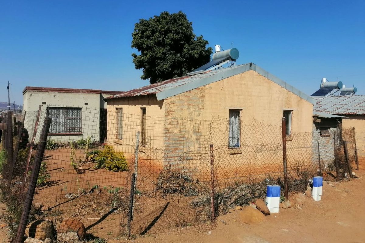Exterior view of a single-story residential dwelling featuring a corrugated metal roof, solar water heating system, and perimeter wire mesh fencing.