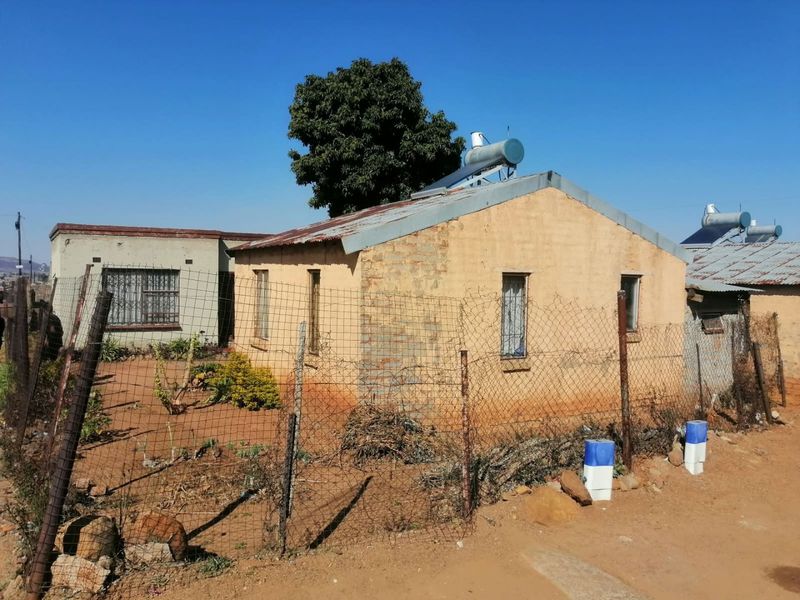 Exterior view of a single-story residential dwelling featuring a corrugated metal roof, solar water heating system, and perimeter wire mesh fencing.