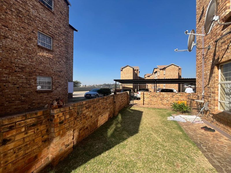 A rear exterior view of an apartment complex featuring a private lawn area, face brick walls, and a communal carport structure.
