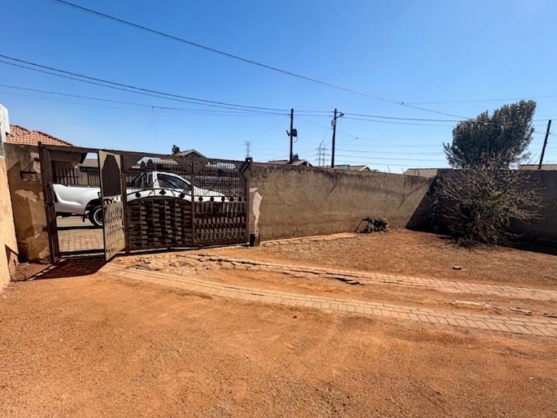 Front exterior view showing a metal driveway gate, perimeter wall, and a paved driveway area in need of maintenance.