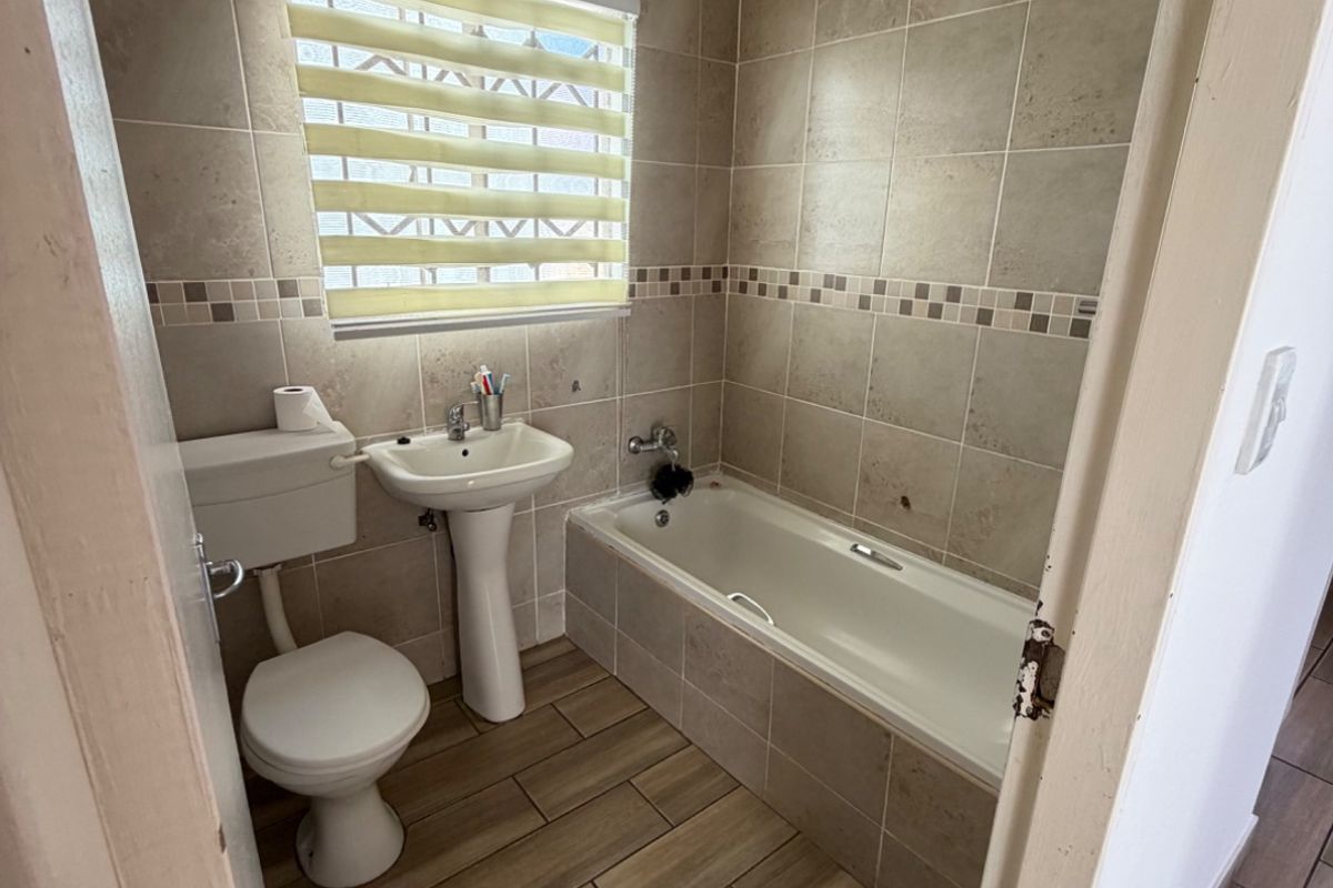 A compact bathroom featuring a pedestal basin, toilet, and bathtub with neutral wall and floor tiling, requiring some maintenance and cosmetic updates.