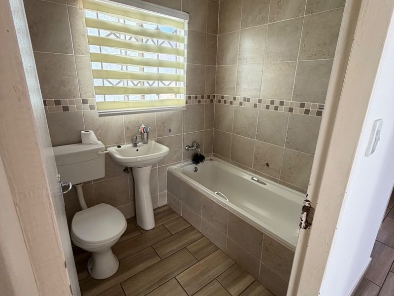 A compact bathroom featuring a pedestal basin, toilet, and bathtub with neutral wall and floor tiling, requiring some maintenance and cosmetic updates.