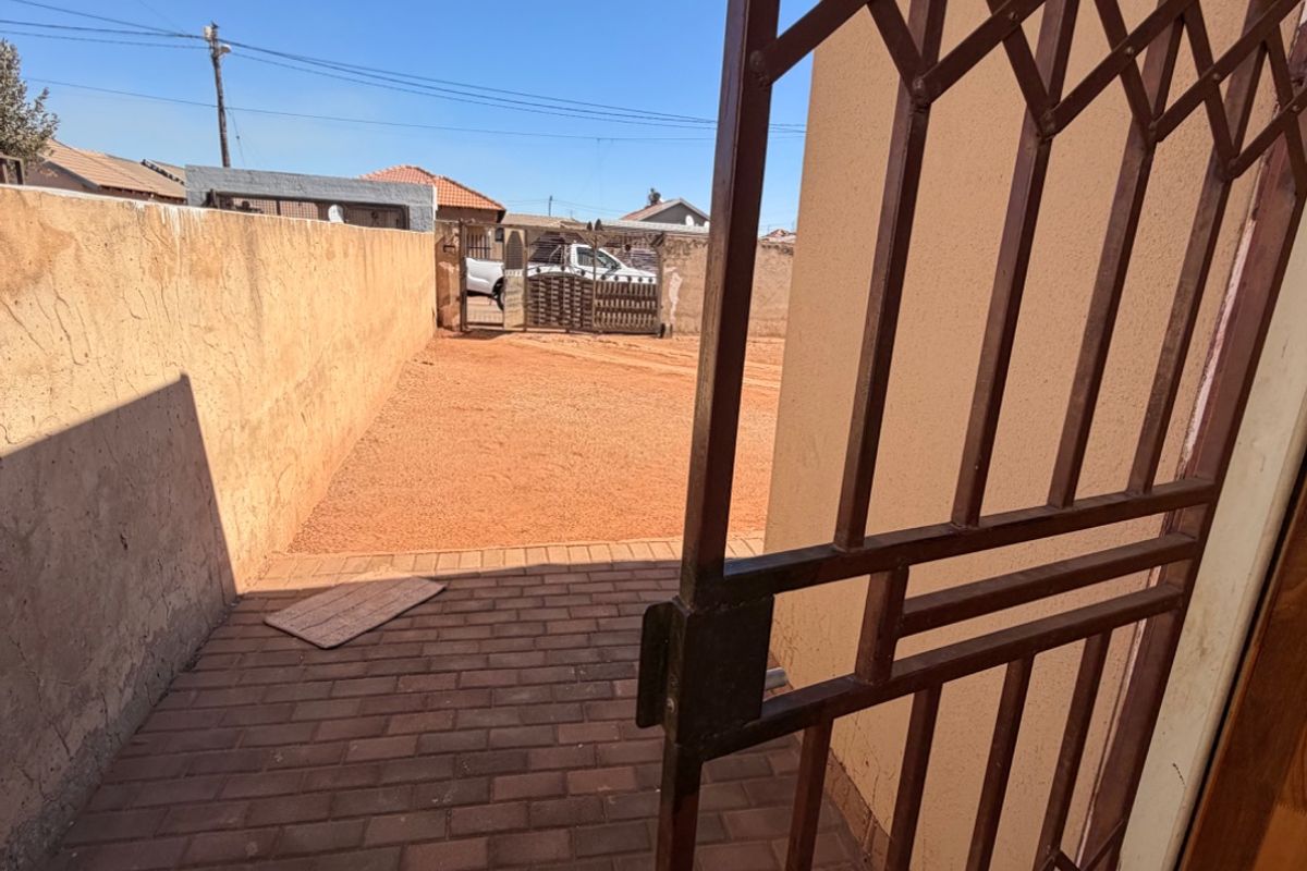 A view of the rear exterior area featuring a paved walkway, security gate, and a boundary wall enclosing the property.