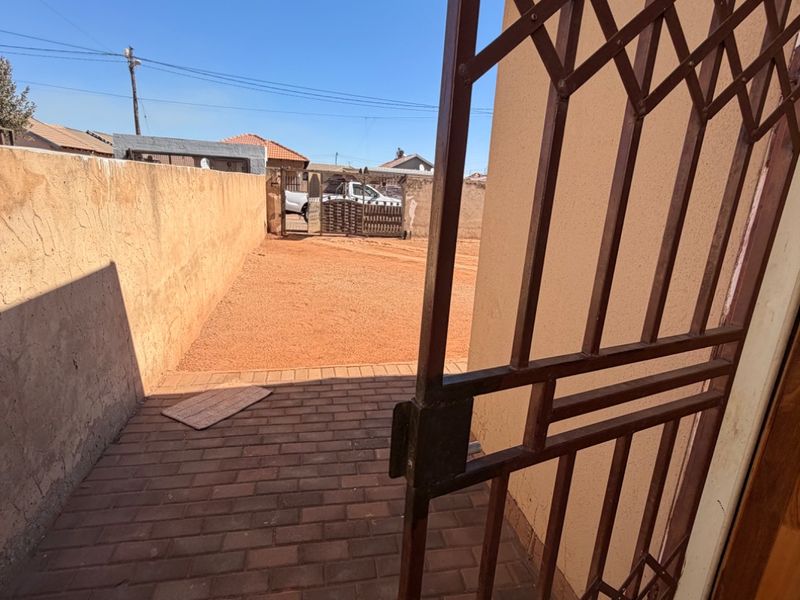 A view of the rear exterior area featuring a paved walkway, security gate, and a boundary wall enclosing the property.