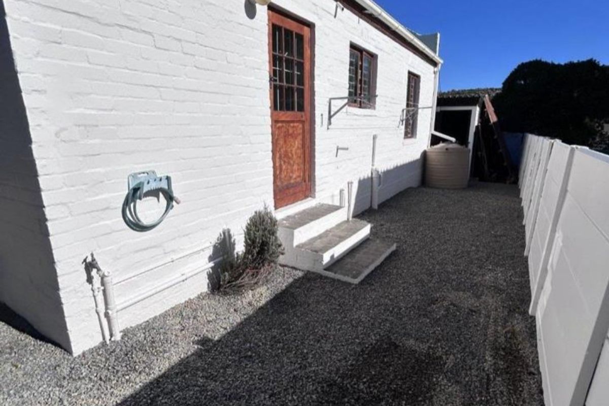 Rear exterior view of the property featuring a painted brick facade, timber door with concrete steps, and a gravel side yard with perimeter walling.