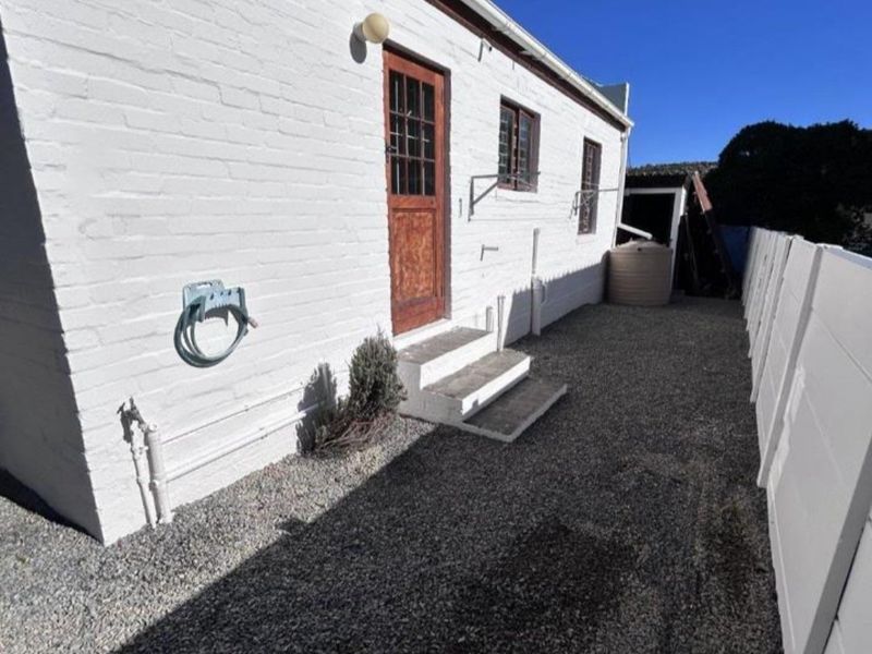 Rear exterior view of the property featuring a painted brick facade, timber door with concrete steps, and a gravel side yard with perimeter walling.