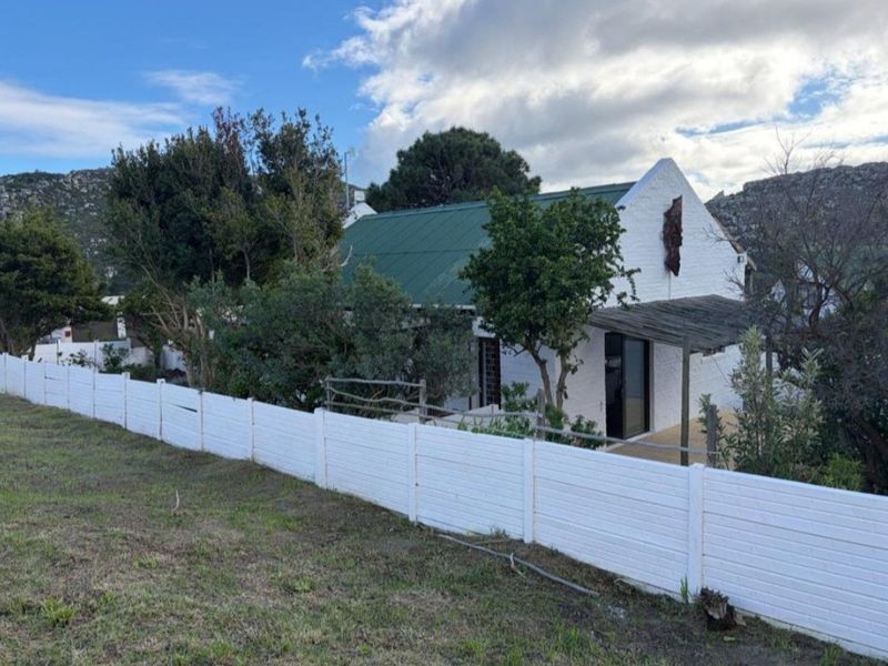 A charming cottage-style home featuring a green metal roof, white brick exterior, and a timber pergola, enclosed by a white boundary fence.