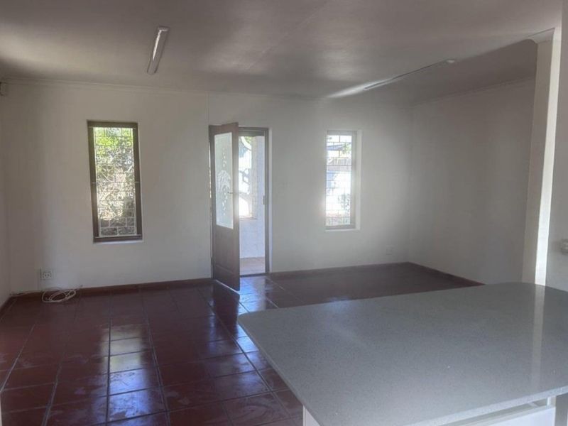 An empty open-plan living area featuring tiled flooring, fluorescent lighting, and a kitchen island, with direct access to the exterior.