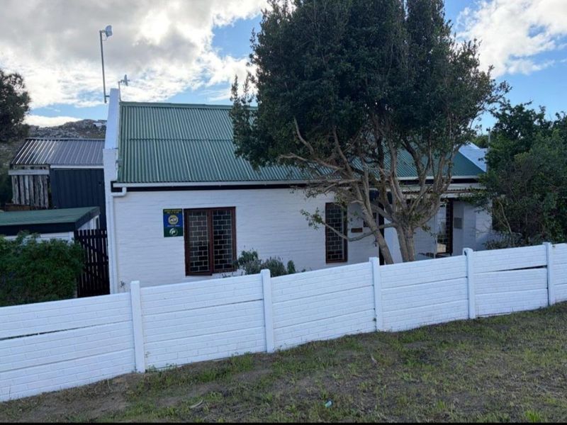 Front exterior view of a cottage-style house featuring a green corrugated metal roof, white painted brick facade, and a secure boundary wall.