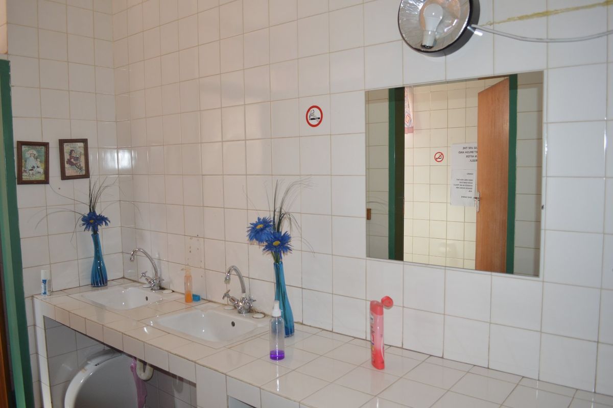 Commercial bathroom featuring a tiled vanity with dual sinks, wall-mounted mirror, and ceramic wall tiling. Requires maintenance on fixtures and wall finishes.