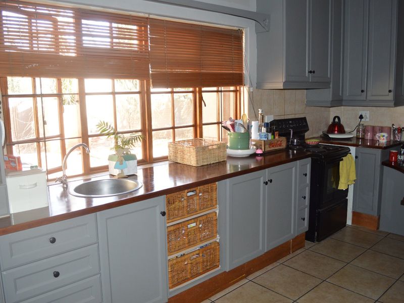 A functional kitchen featuring grey cabinetry, wooden countertops, a stainless steel sink, and tiled flooring with natural light from large timber-framed windows.