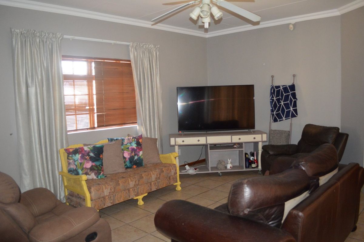 A comfortable living room featuring tiled flooring, a ceiling fan, and large windows fitted with venetian blinds and curtains.