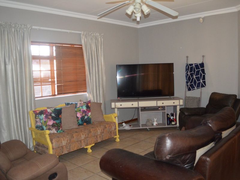 A comfortable living room featuring tiled flooring, a ceiling fan, and large windows fitted with venetian blinds and curtains.